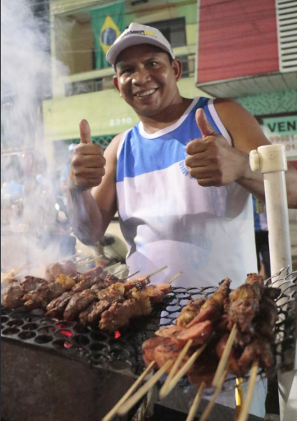 Aluno sorrindo atrás da churrasqueira cheia de espetinhos